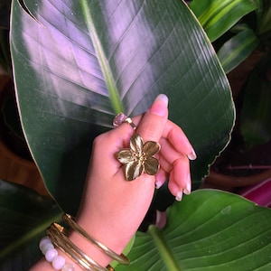May include: A gold flower ring with a delicate design. The ring is worn on a hand with multiple gold bangles and a white beaded bracelet.