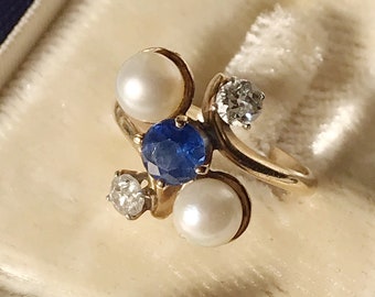 Könnte beinhalten: Ein goldener Ring mit einem blauen Saphir und drei weißen Perlen. Der Ring befindet sich in einer Samt-Ringbox.