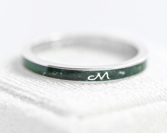 Könnte beinhalten: Ein silberner Ring mit einem dunkelgrünen Band, auf dem der Buchstabe "M" in zarter Schrift steht. Der Ring ist vor einem weichen, weißen Hintergrund platziert, der sein elegantes Design hervorhebt. Ein minimalistisches Schmuckstück.