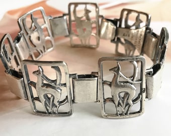 May include: A silver bracelet featuring a repeating pattern of stylised deer figures. The bracelet is made of linked rectangular pieces with cut-out designs.