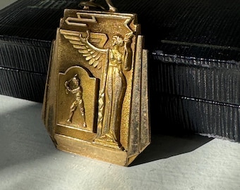Könnte beinhalten: Ein goldfarbener Metallanhänger mit einer geflügelten Figur, die eine Fackel hält, und einem Baseballspieler. Der Anhänger befindet sich in einer schwarzen Schachtel.