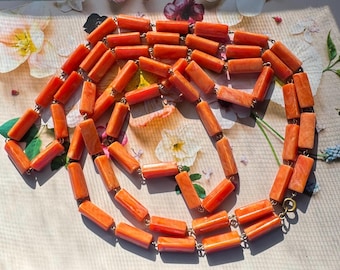 Könnte beinhalten: Eine lange Halskette, die aus zahlreichen zylindrischen, orangefarbenen Perlen besteht, die in einer durchgehenden Schlaufe aufgereiht sind. Die Perlen haben eine glatte, polierte Oberfläche mit subtilen Farbnuancen und sind durch kleine Metallglieder verbunden.