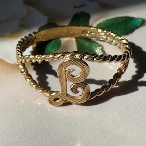 May include: A gold-toned ring featuring a twisted rope-style band and a central, stylized letter L. The letter has a textured, etched surface finish, and the band is constructed from two parallel twisted wires that meet at the letter centerpiece.
