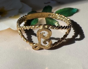 Könnte beinhalten: Ein goldfarbener Ring mit einem gedrehten, seilartigen Band und einem zentralen, stilisierten Buchstaben L. Der Buchstabe hat eine texturierte, geätzte Oberfläche, und das Band besteht aus zwei parallelen, gedrehten Drähten, die sich am Mittelstück treffen.