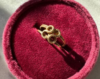 Puede incluir: Un anillo de oro con un diseño de nudo, presentado en una caja de anillo de terciopelo rojo.