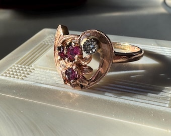 Könnte beinhalten: Ein goldener Ring mit einem herzförmigen Design, das mit einem Cluster aus rosafarbenen Edelsteinen und einem einzigen klaren Edelstein in der Mitte verziert ist.