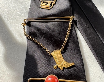 Könnte beinhalten: Drei goldene Krawattenklammern mit Ketten. Die obere Klammer hat ein geometrisches Design, die mittlere Klammer hat ein Stiefeldesign und die untere Klammer hat einen roten Stein.
