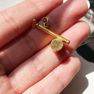May include: A gold-toned pendant necklace featuring a small, round charm with a long, thin bar attached. The charm is smooth and has a slightly textured surface.