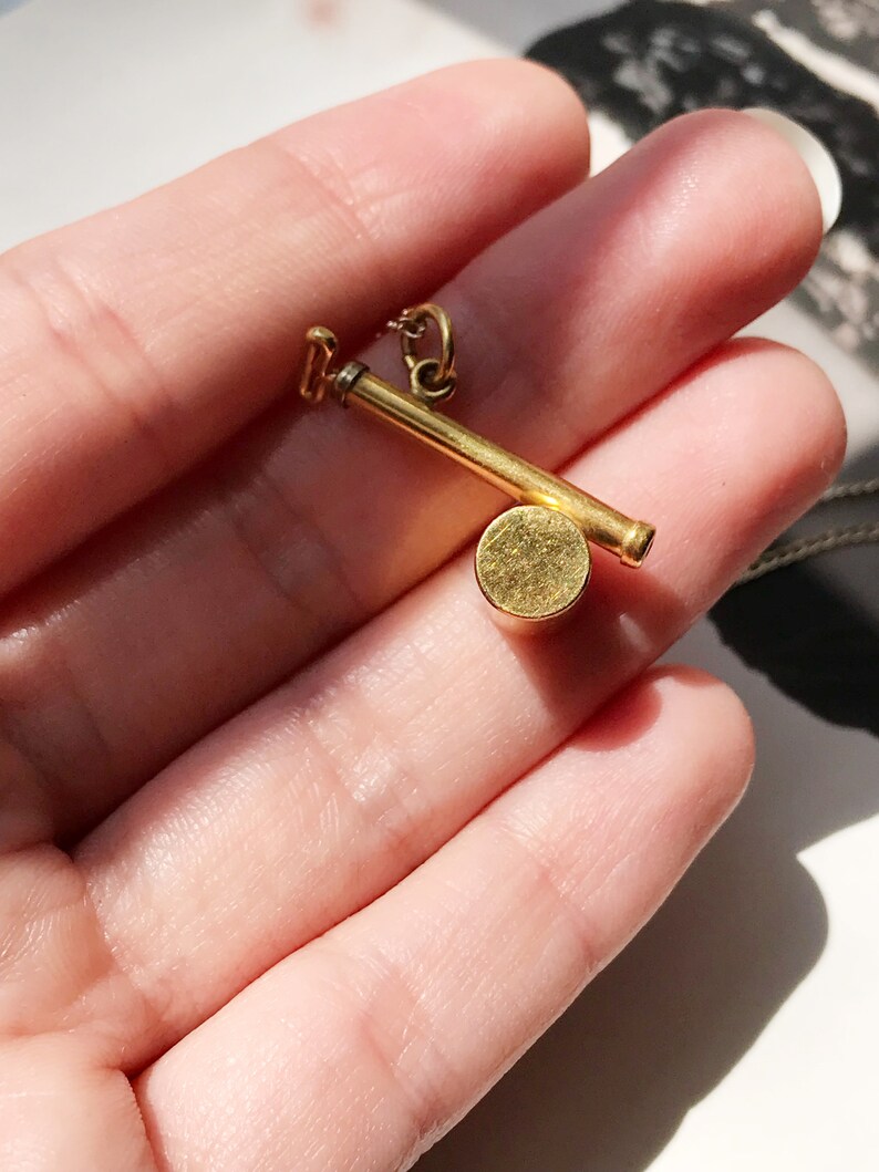 May include: A gold-toned pendant necklace featuring a small, round charm with a long, thin bar attached. The charm is smooth and has a slightly textured surface.