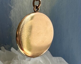 Könnte beinhalten: Ein runder, glatter goldfarbener Medaillon-Anhänger, der an einer passenden goldfarbenen Metallkette hängt. Das Medaillon hat eine polierte, reflektierende Oberfläche mit einem einfachen kreisförmigen Design und einem kleinen Verbindungsring.