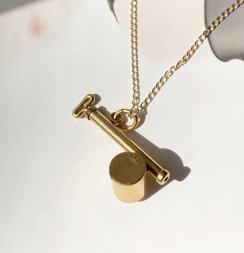 May include: A gold chain necklace with a pendant that looks like a small, cylindrical key. The key has a small, round head and a long, thin shaft.