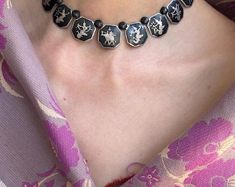 Könnte beinhalten: Silberfarbene Halskette mit schwarzen achteckigen Anhängern, die silberne Figuren zeigen. Die Halskette wird vor dem Hintergrund eines lila-rosa Schals mit Blumenmuster getragen. Die Kette ist filigran und kleine schwarze Perlen verbinden die Anhänger.