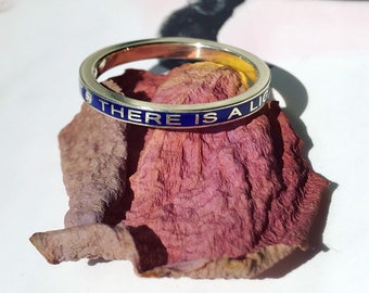 Könnte beinhalten: Ein dünner Silberring mit einem blauen Emailleband und der Aufschrift "THERE IS A LIGHT" in Weiß. Der Ring liegt auf einem rosafarbenen Rosenblatt.