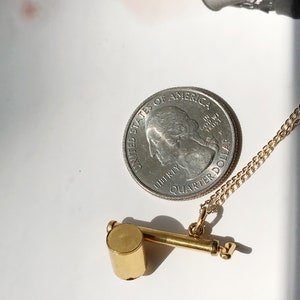 May include: A gold whistle necklace with a silver quarter dollar coin charm. The coin has the words "United States of America" and "In God We Trust" on it. The necklace is on a white surface.