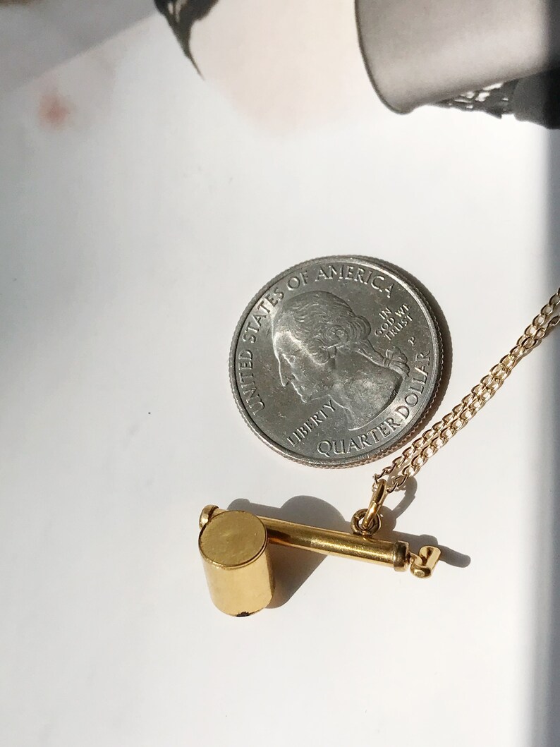 May include: A gold whistle necklace with a silver quarter dollar coin charm. The coin has the words "United States of America" and "In God We Trust" on it. The necklace is on a white surface.