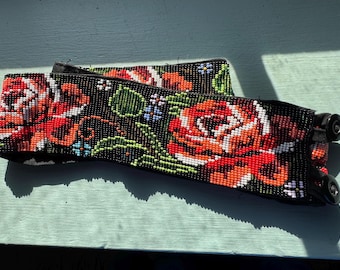 Könnte beinhalten: Ein breiter Gürtel mit aufwendigen Perlenstickereien auf schwarzem Grund. Das Design zeigt große, leuchtend rote und orangefarbene Rosen mit grünen Blättern sowie kleine blaue und weiße Blumenakzente. Der Gürtel hat zwei schwarze Druckknöpfe.