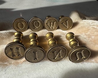 Könnte beinhalten: Acht Messing-Buchstabenstempel mit verschiedenen Buchstaben, darunter H, I, N und U. Die Stempel sind in zwei Reihen auf einer weißen Oberfläche angeordnet.