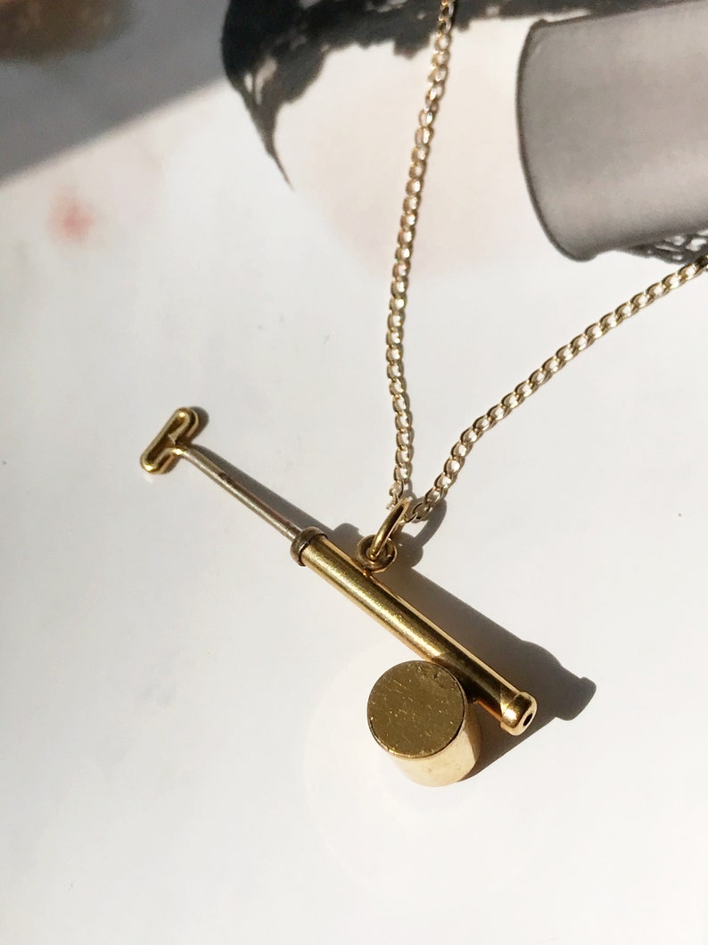 May include: A gold necklace with a pendant shaped like a small, handheld pump. The pump has a round base and a long, thin handle.
