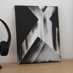 May include: Abstract black and white painting on canvas with a geometric design. The painting features a white X shape with black lines and shapes around it.