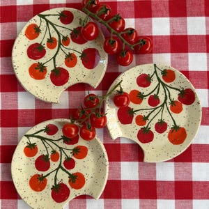 Puede incluir: Tres platos de cerámica blanquecina con diseño de tomate, cada uno con un recorte único. Los platos presentan tomates rojos y naranjas sobre tallos verdes, con un acabado moteado. Los tomates cherry frescos están dispuestos cerca sobre un mantel a cuadros rojos y blancos.