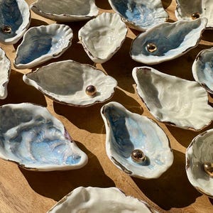 Puede incluir: Una colección de platos de cerámica en forma de concha de ostra con detalles dorados. Cada plato presenta una perla en el centro.