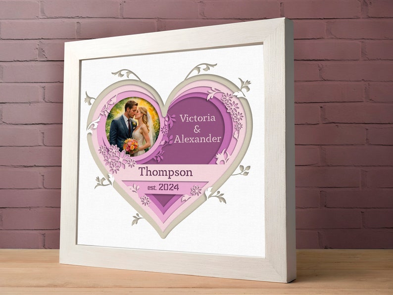 K&ouml;nnte beinhalten: Ein wei&szlig;er Rahmen mit einem mehrschichtigen Herzdesign in Rosa und Grau. Das Herz zeigt im Zentrum ein Foto eines Paares, darunter der Text "Victoria & Alexander Thompson est. 2024".