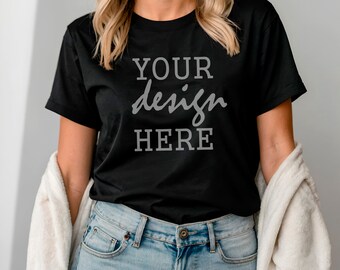 Puede incluir: Una camiseta negra con el texto "YOUR design HERE" impreso en la parte delantera. La camiseta la lleva puesta una persona con el pelo largo y rubio y va combinada con unos vaqueros azules.