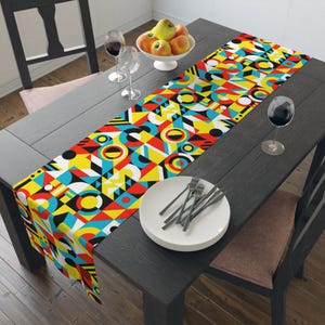 May include: A colourful geometric table runner with red, yellow, blue and black shapes. The runner is on a dark wood table with a white plate and cutlery.