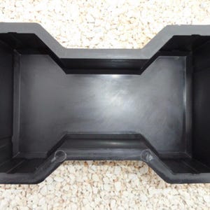 May include: A black plastic paving stone mould. The mould has a distinctive shape with a central rectangular area and an hourglass-like design. It is designed to create durable and decorative paving stones for outdoor use, such as patios or walkways.