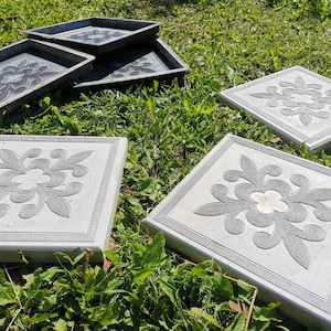 Puede incluir: Varias losas de jardín de hormigón gris con un diseño floral dispuestas sobre hierba verde. Las losas tienen forma cuadrada con un borde decorativo. Algunas son planas, otras en forma de bandeja.