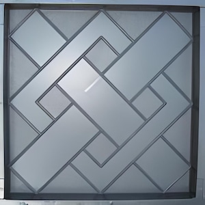 May include: A square, geometric design featuring a pattern of overlapping rectangles and squares in shades of gray. The design is framed by a dark gray border, creating a modern, abstract aesthetic. The glass-like material reflects light.