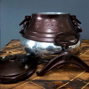 Puede incluir: Una olla de cocina bicolor marrón y plateada con tapa a juego y un trípode. La olla tiene una banda decorativa alrededor del centro y asas a los lados. La tapa está a un lado, revelando el interior.