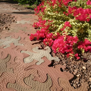 May include: Interlocking outdoor pavers in a repeating lizard or reptile shape, featuring alternating terracotta and light tan colors with textured surfaces, arranged in a puzzle-like pattern alongside a cluster of vibrant pink flowers.