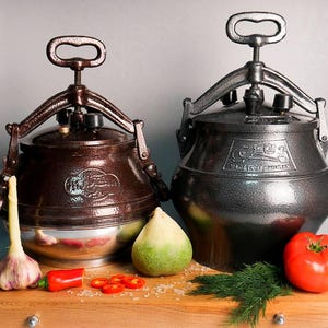 以下が含まれることがあります： 茶色とグレーの金属製圧力鍋2つ。ハンドルとロック機構が付いています。鍋は、トマト、ニンニク、唐辛子などの野菜が置かれた木製の表面に置かれています。