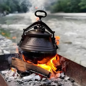 May include: A black cast iron cooking pot with a handle and pressure release valve is heated over a wood fire. The pot has decorative floral and script details. Flames and smoke rise from the burning wood.