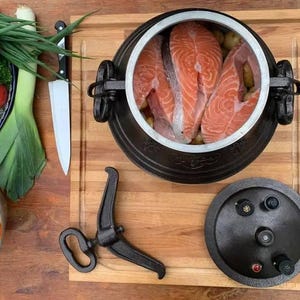 Puede incluir: Vista aérea de una olla de hierro fundido con filetes de salmón y patatas. Una tabla de cortar de madera, un cuchillo y verduras frescas están dispuestos alrededor de la olla. La olla tiene una tapa y un soporte.