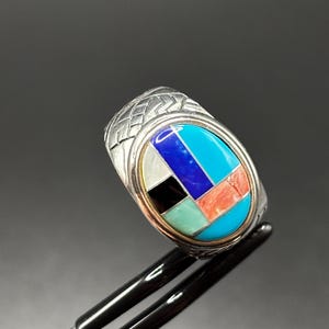 May include: Silver ring with an oval inlay. The inlay features a geometric design in turquoise, blue, black, white, and coral. The ring has a textured band and a gold border around the inlay.