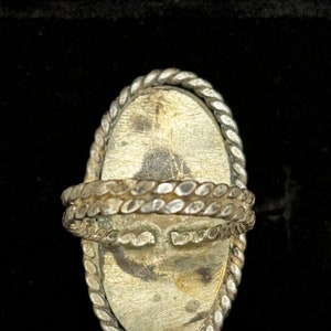 May include: A silver ring with a large oval-shaped stone set in a braided metal bezel. The ring is designed to be worn on the finger.