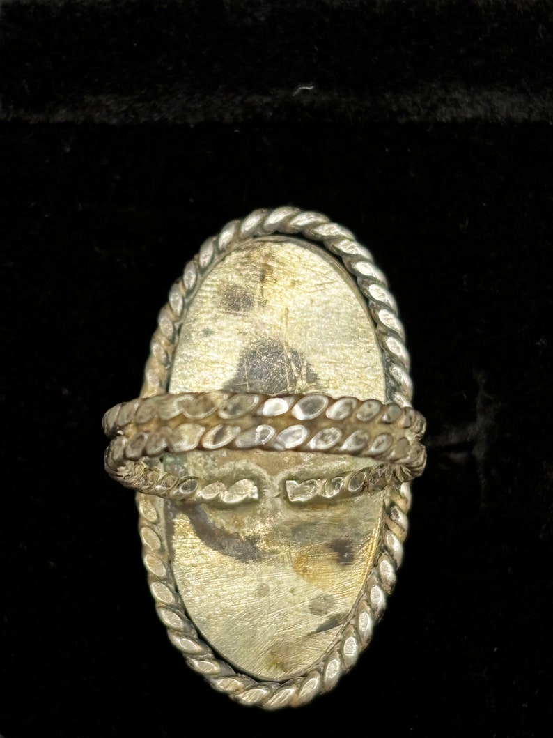 May include: A silver ring with a large oval-shaped stone set in a braided metal bezel. The ring is designed to be worn on the finger.