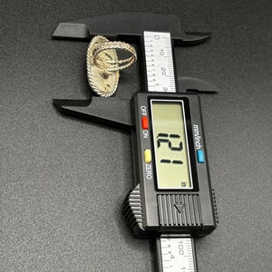 May include: A silver ring with a textured design is measured with a digital caliper. The caliper reads 121 inches.