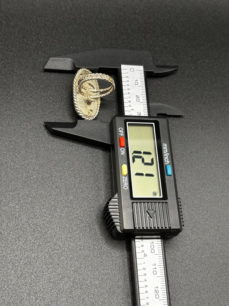May include: A silver ring with a textured design is measured with a digital caliper. The caliper reads 121 inches.