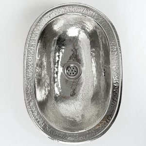 May include: A silver oval-shaped bathroom sink with a hammered finish and a drain in the centre.