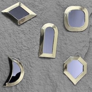 Puede incluir: Cinco pequeños espejos decorativos con marcos de latón y superficies reflectantes negras. Los espejos tienen varias formas geométricas, incluyendo un diamante, un cuadrado, un cuadrado redondeado, un arco y un octágono.