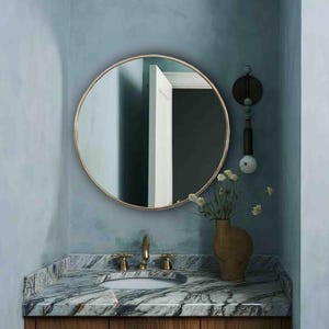 May include: A bathroom vanity with a marble countertop and wooden cabinets. A round mirror with a gold frame hangs above the sink. A vase with flowers sits on the countertop.