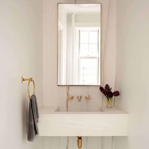 以下が含まれることがあります： 長方形の白い洗面台、金色の金具、金色のフレームの鏡を備えたモダンなバスルームの洗面化粧台。灰色のタオルが金色のタオルリングに掛かっています。濃い赤色の花の小さな花瓶がカウンターに置かれています。