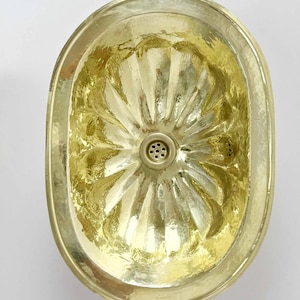 May include: A gold oval-shaped bathroom sink with a decorative, sunburst-like pattern. The sink has a single drain hole in the centre.