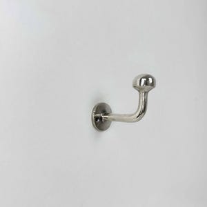 Puede incluir: Un gancho de pared plateado con una cabeza redondeada y un brazo curvado. El gancho está montado sobre una base circular y está diseñado para colgar objetos. El gancho es de metal y está fijado a una pared blanca.