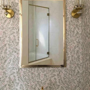 Puede incluir: Interior de baño con espejo con marco dorado, puerta de ducha de cristal y grifos dorados. Las paredes están cubiertas con papel pintado con un patrón de hojas verdes y bayas rosas. Dos apliques dorados con pantallas de cristal flanquean el espejo.