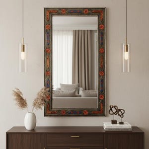 Op de afbeelding: Een rechthoekige spiegel met een decoratieve lijst met rode, blauwe en oranje bloemmotieven. De spiegel hangt boven een donkerbruin houten dressoir met drie lades en een deur. Twee hanglampen met cilindrische glazen kappen flankeren de spiegel.