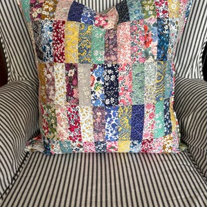 May include: A square patchwork cushion featuring a mix of floral fabrics in pinks, blues, yellows, and greens. The cushion is set on a striped chair, offering a vibrant and decorative touch for home interiors.